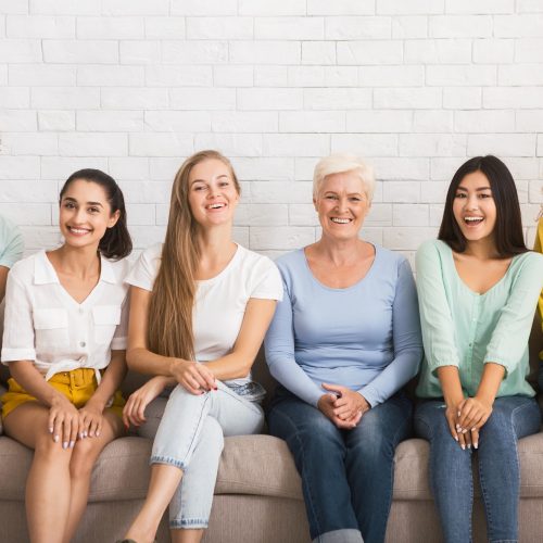 Smiling Diverse Women Sitting On Sofa Over White Brick Wall
